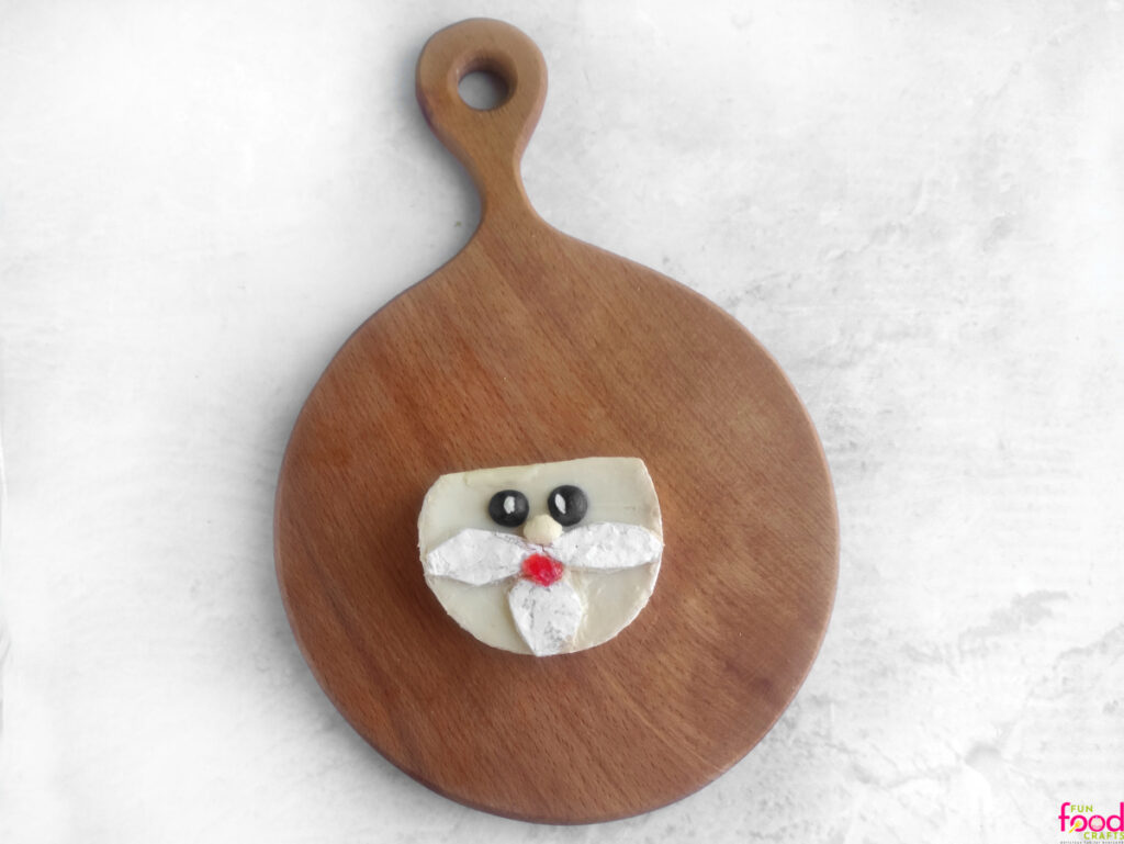 Overhead view of a Brie cheese round decorated with blueberries, mayo, cheese rind, and a red candy to look like Uncle Sam’s face on a wooden cutting board.
