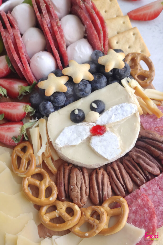 Close-up of Uncle Sam’s cheese face with blueberry eyes, cherry candy mouth, white cheese mustache, braided cheese curls, and star crackers on a bed of blueberries for his hat.