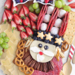 Overhead view of a full Uncle Sam Charcuterie Board with red, white, and blue candies, strawberries, grapes, cheese stars, salami, and a Brie cheese Uncle Sam face in the center.