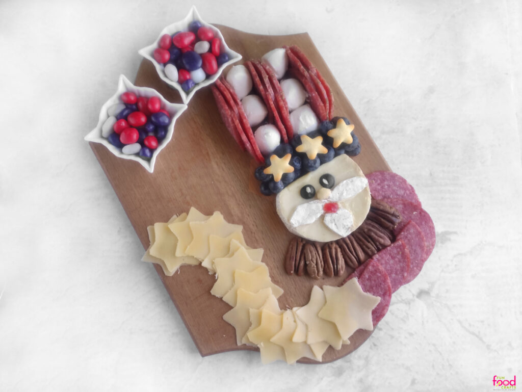 Partially assembled Uncle Sam Charcuterie Board with cheese face, blueberry and salami hat, pecan bowtie, and candy bowls on a wooden board.