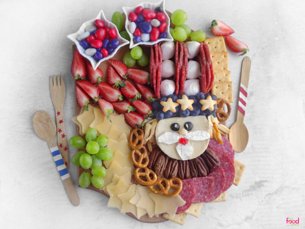 Fully assembled 4th of July Uncle Sam Charcuterie Board surrounded by strawberries, grapes, cheese stars, patriotic crackers, candies, and festive wooden spoons.