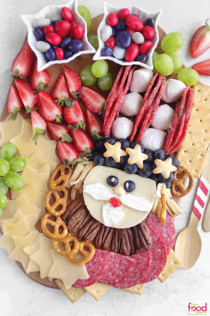 Overhead view of a full Uncle Sam Charcuterie Board with red, white, and blue candies, strawberries, grapes, cheese stars, salami, and a Brie cheese Uncle Sam face in the center.