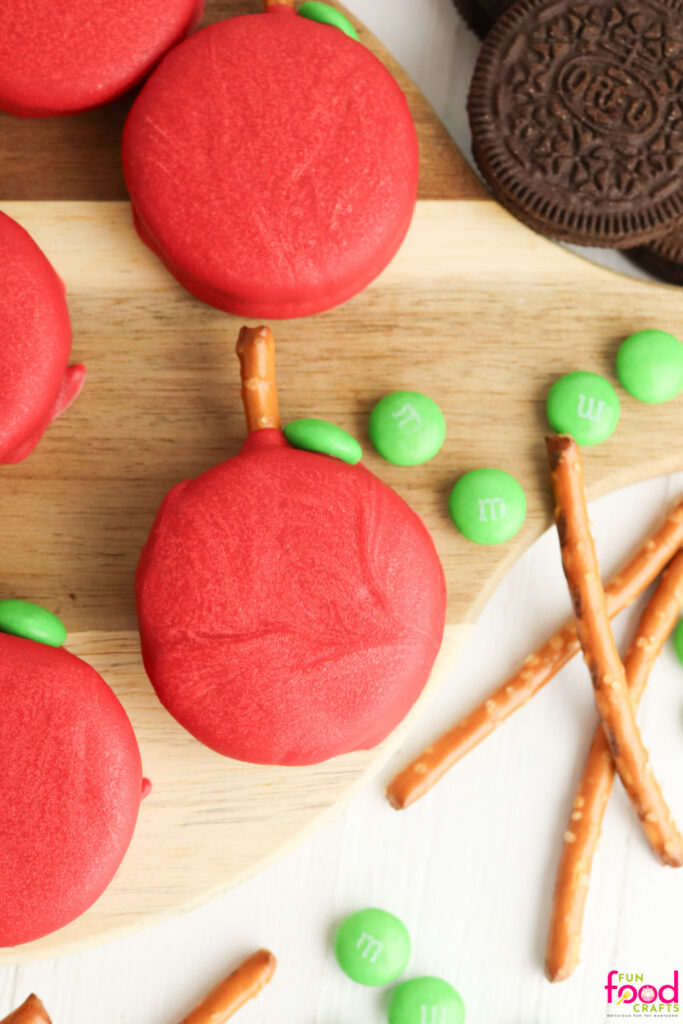 Red chocolate-covered Oreos made to look like apples using green candies and pretzel sticks