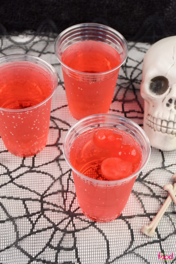 Close-up of red Halloween drinks with cherry skull-shaped ice cubes melting inside plastic cups beside a skull prop and faux bones.