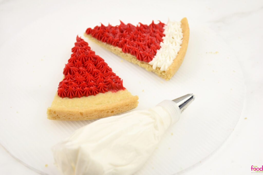 Two cookie slices decorated as Santa hats with red star-tip piping and a fluffy white brim, icing bag and tip beside them.