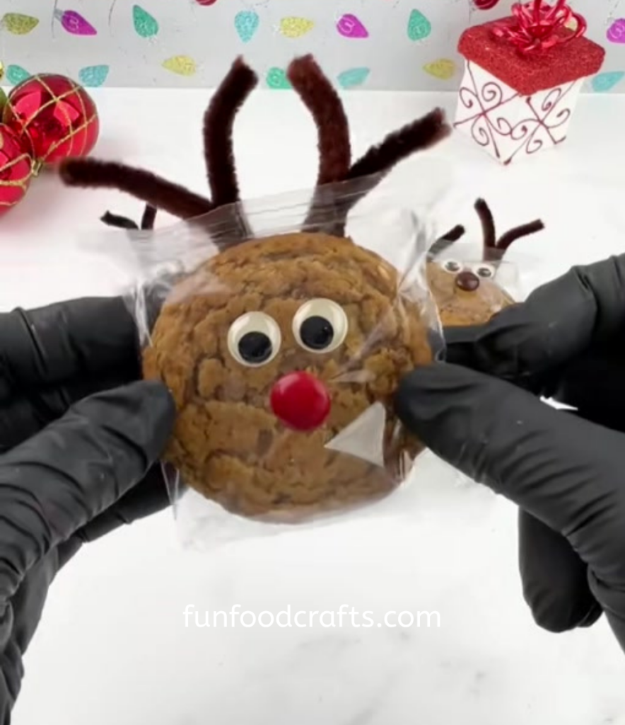 Hand holding a finished oatmeal creme pie reindeer with googly eyes, brown pipe cleaner antlers, and a red M&M nose attached to the wrapper.